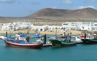 Fishing boats docked at Caleta de Sebo harbor, La Graciosa, Canary Islands, with white buildings and hills in the background.