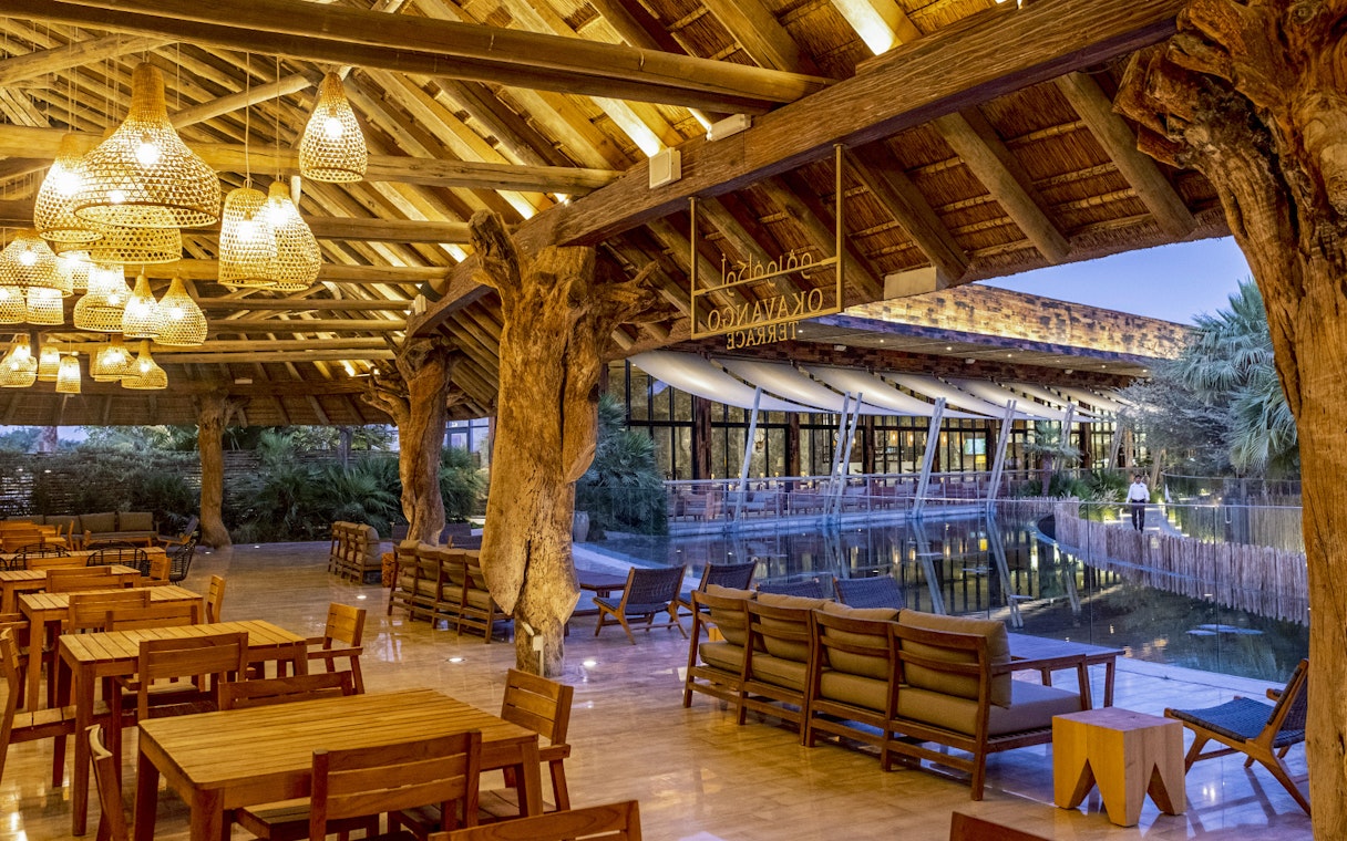 Outdoor dining area at Okavango Terrace with wooden tables and chairs under a thatched roof.