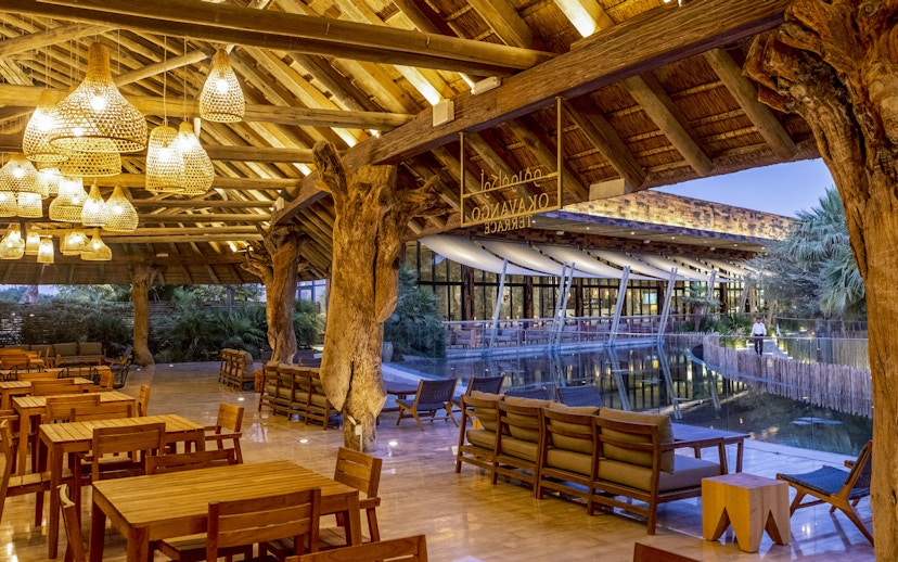 Outdoor dining area at Okavango Terrace with wooden tables and chairs under a thatched roof.