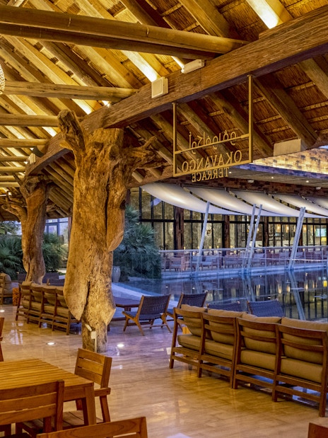 Outdoor dining area at Okavango Terrace with wooden tables and chairs under a thatched roof.