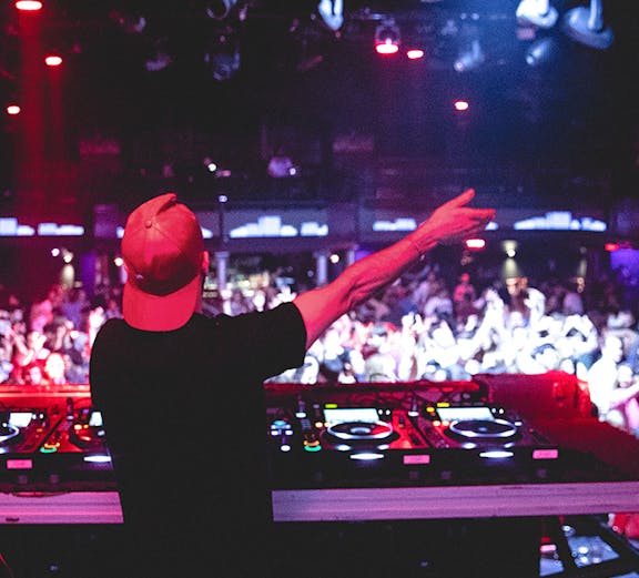DJ performing at nightclub, view from behind with crowd and lights.