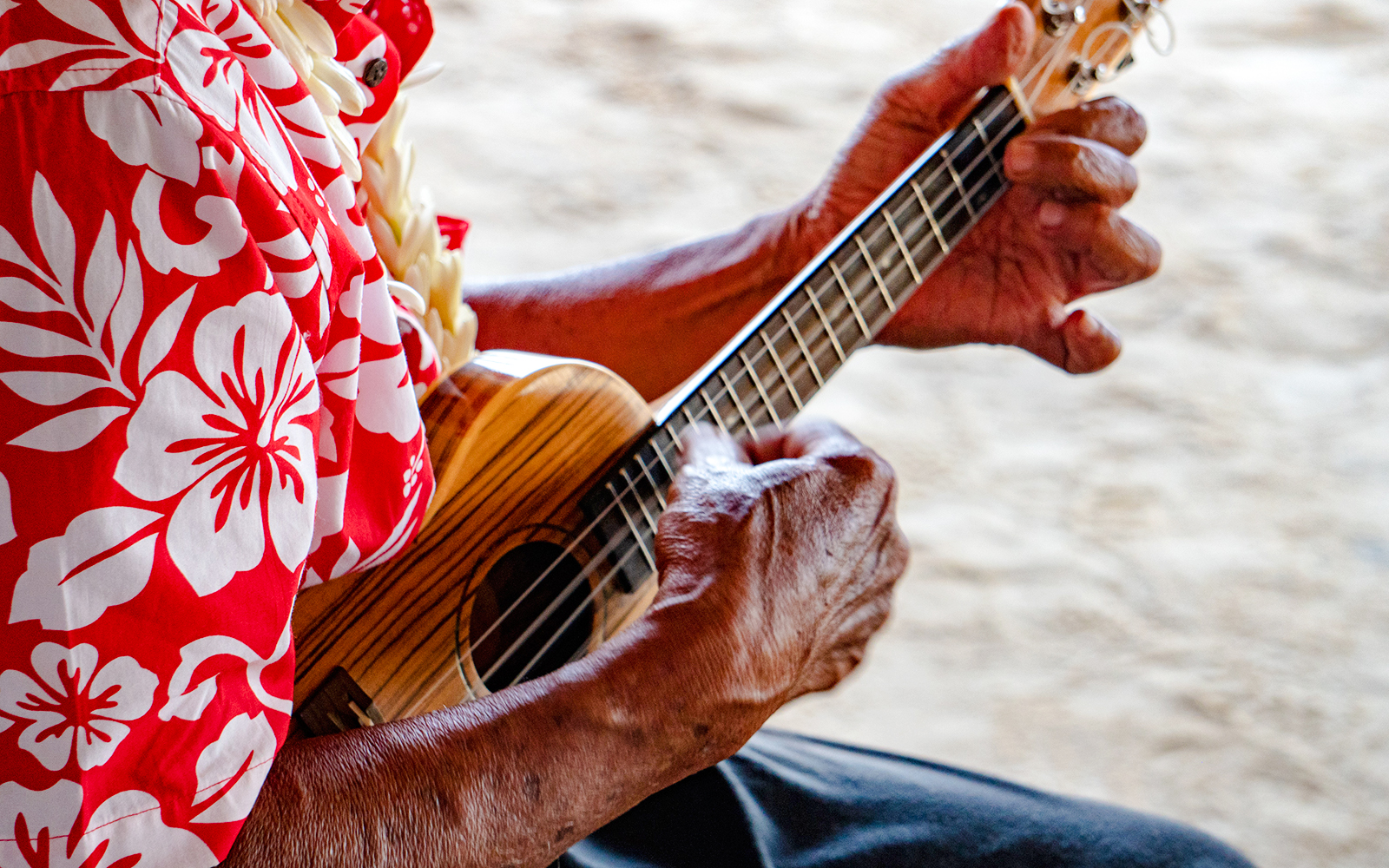 Old man's hands playing a ukulele, wearing a floral shirt.