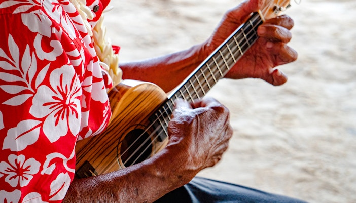 Old man's hands playing a ukulele, wearing a floral shirt.