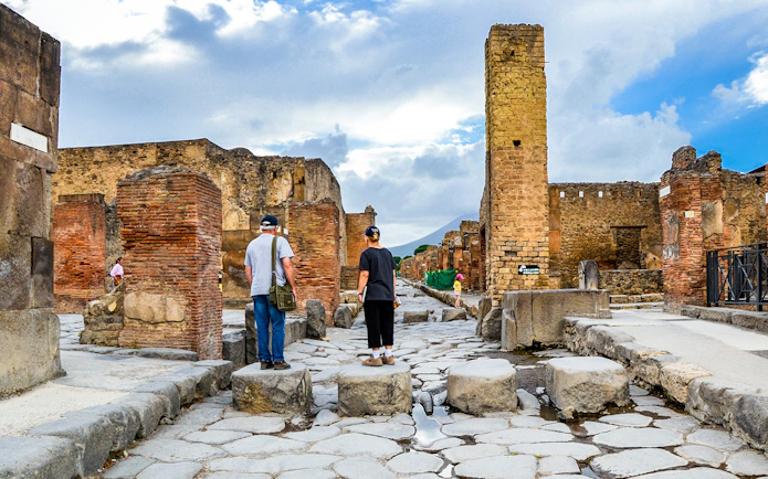 Tourists exploring ancient ruins in Pompeii, Italy, with Mount Vesuvius in the background.