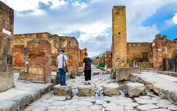 Tourists exploring ancient ruins in Pompeii, Italy, with Mount Vesuvius in the background.