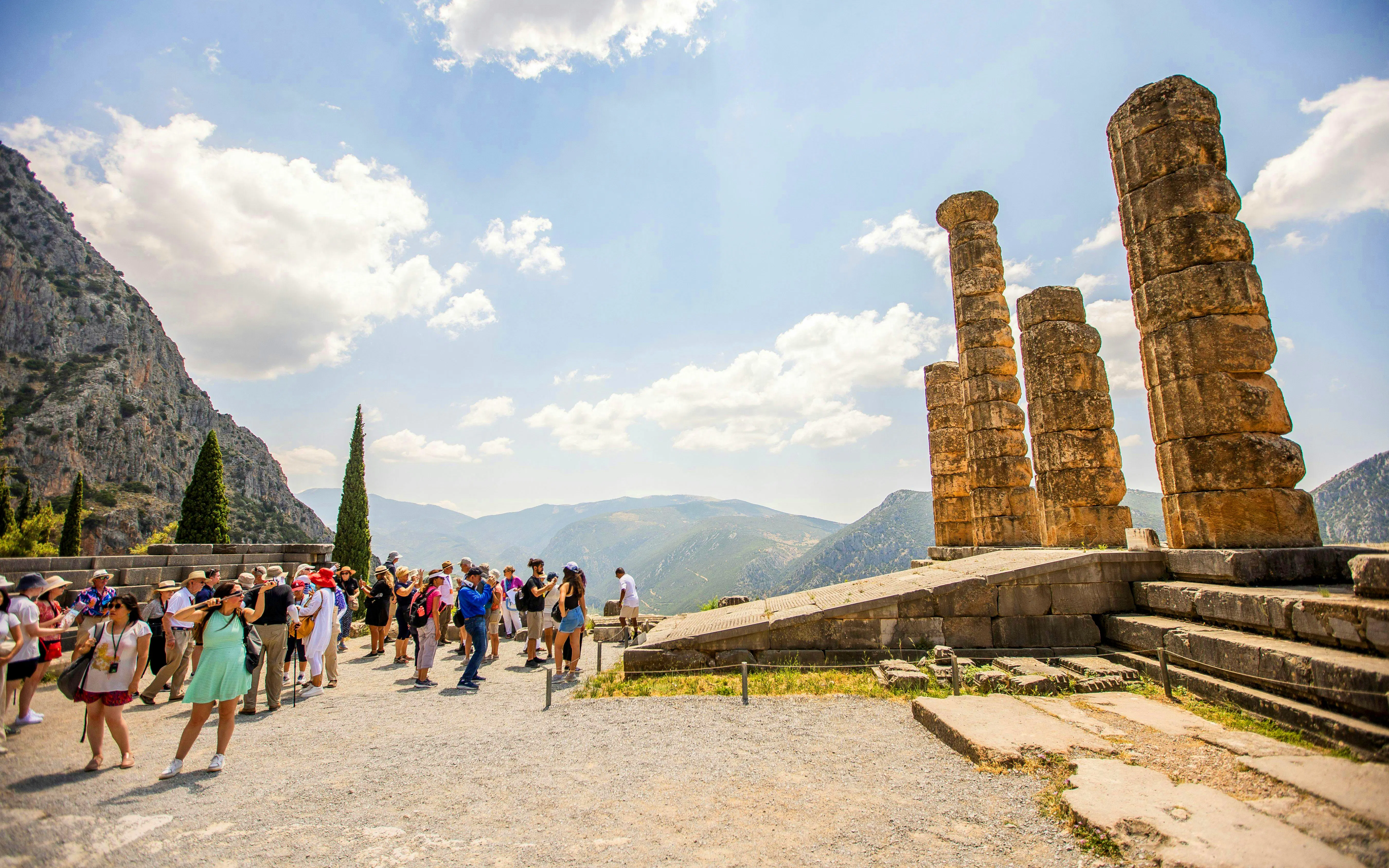 Tourists exploring ancient ruins at Delphi, Greece, with mountainous landscape in the background.