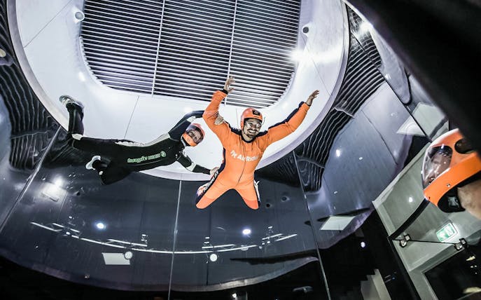 Indoor skydiving with instructor guiding participant in wind tunnel.