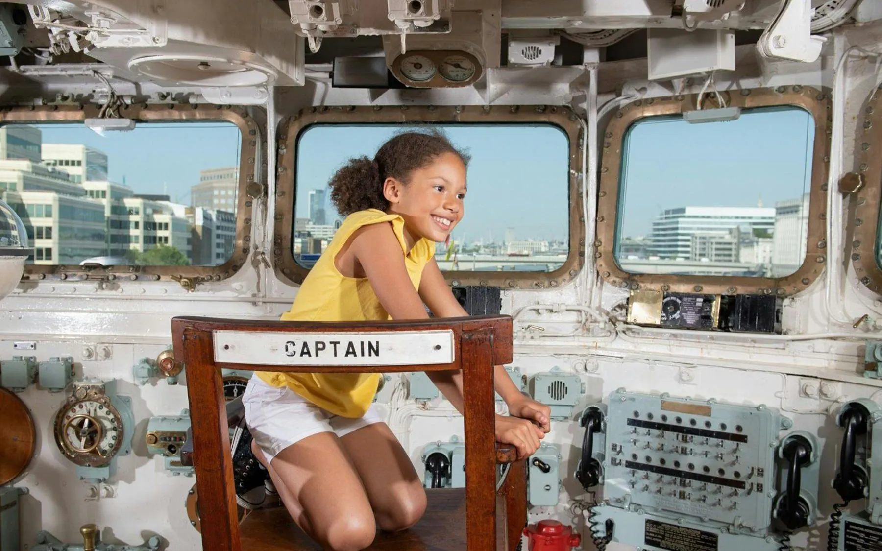 Child sitting in the captain's chair on HMS Belfast with cityscape view.