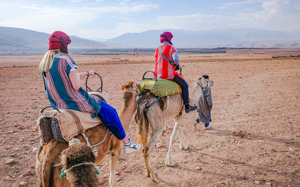 People riding camels in Agafay Desert, Morocco with mountains in the background.