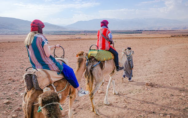 People riding camels in Agafay Desert, Morocco with mountains in the background.