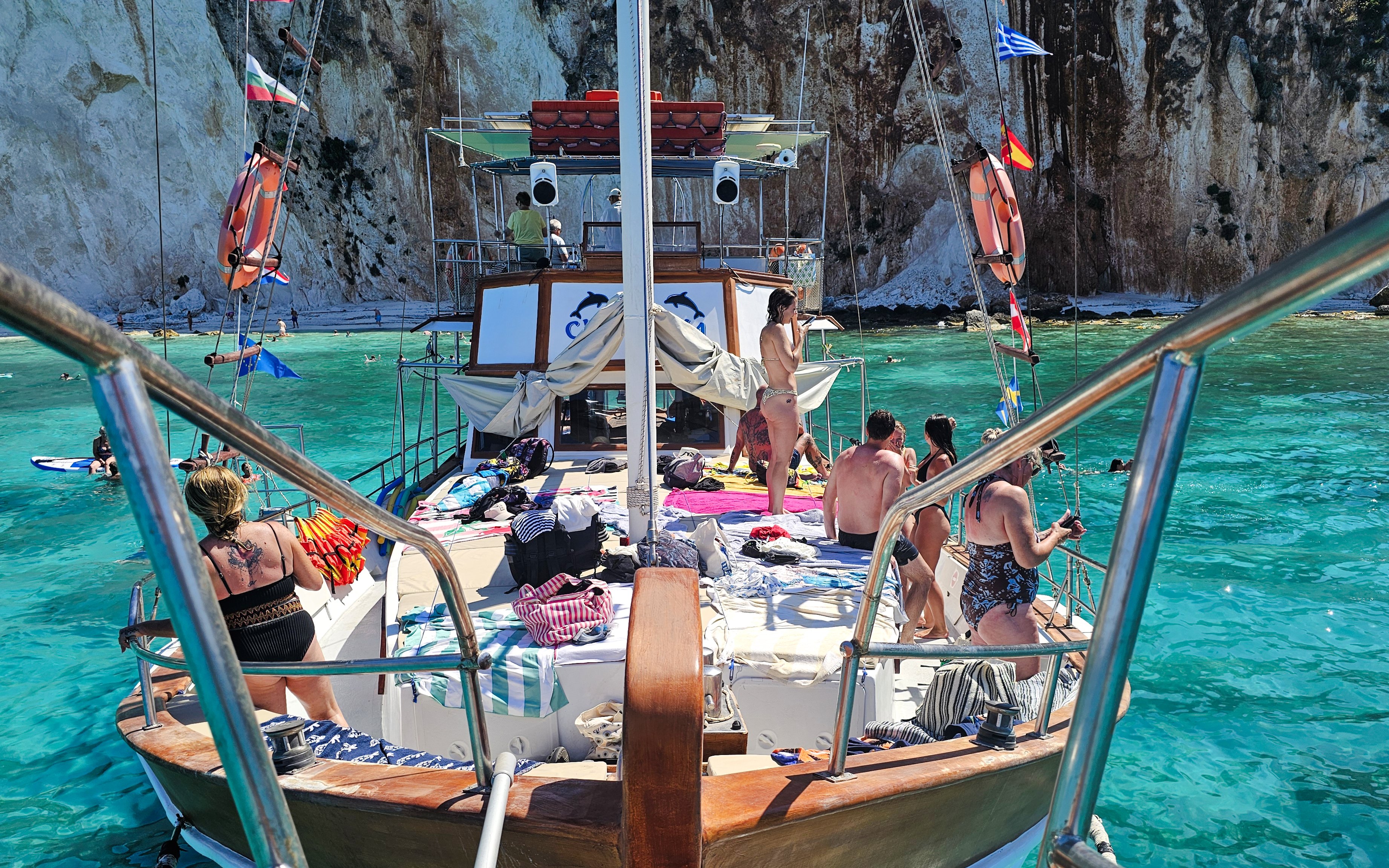 Cruise ship deck with people sunbathing and swimming in turquoise waters near Kefalonia cliffs.