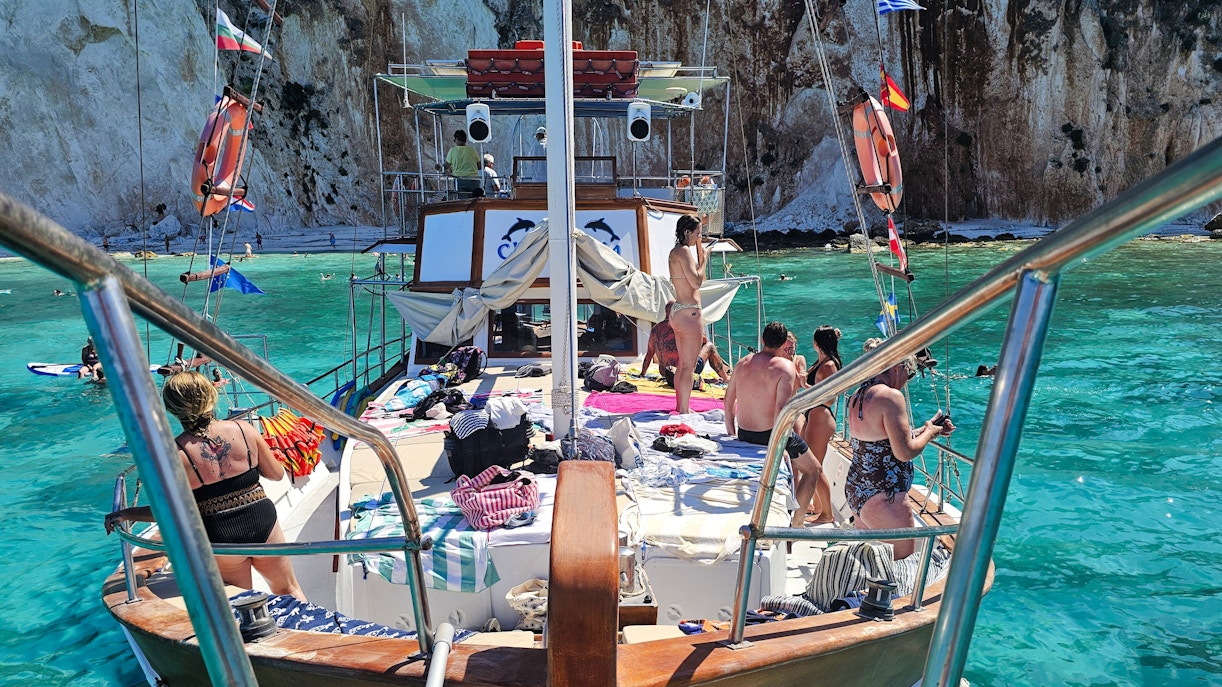 Cruise ship deck with people sunbathing and swimming in turquoise waters near Kefalonia cliffs.