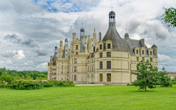 Château de Chambord with lush green grounds under a cloudy sky, part of Chambord and Chenonceau Tour.