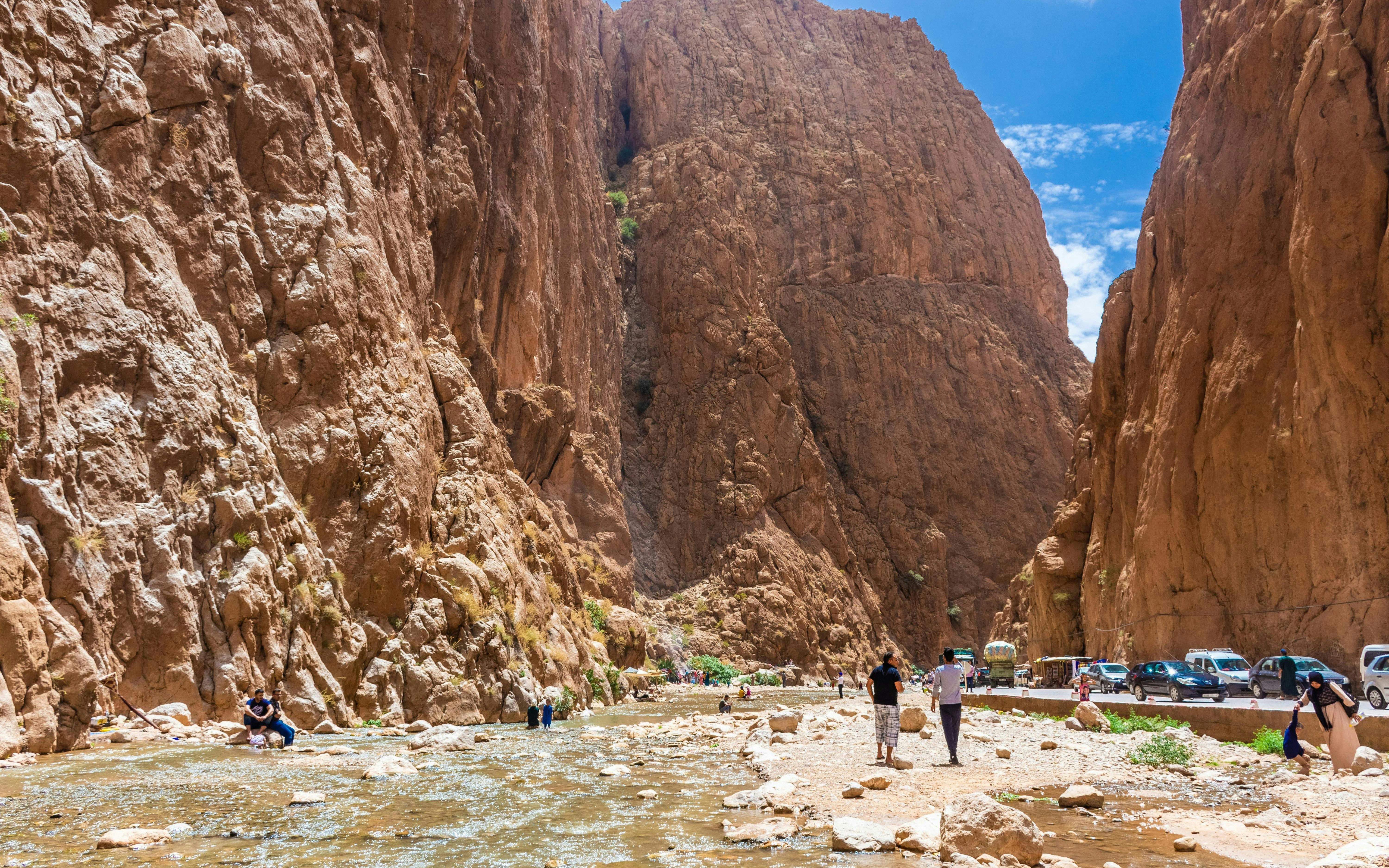Tourists walking along the Todra Gorge in Tinerhir, Morocco, with towering rock cliffs and a shallow river.