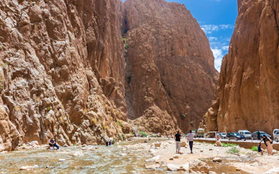 Tourists walking along the Todra Gorge in Tinerhir, Morocco, with towering rock cliffs and a shallow river.