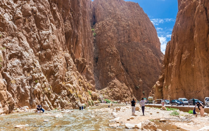 Tourists walking along the Todra Gorge in Tinerhir, Morocco, with towering rock cliffs and a shallow river.