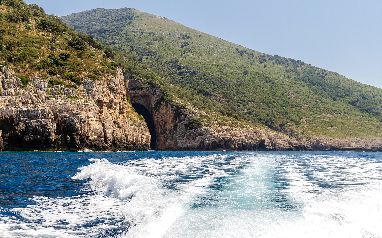 Boat approaching the entrance of Haxhi Ali Cave, surrounded by rocky cliffs and greenery.