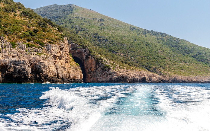 Boat approaching the entrance of Haxhi Ali Cave, surrounded by rocky cliffs and greenery.
