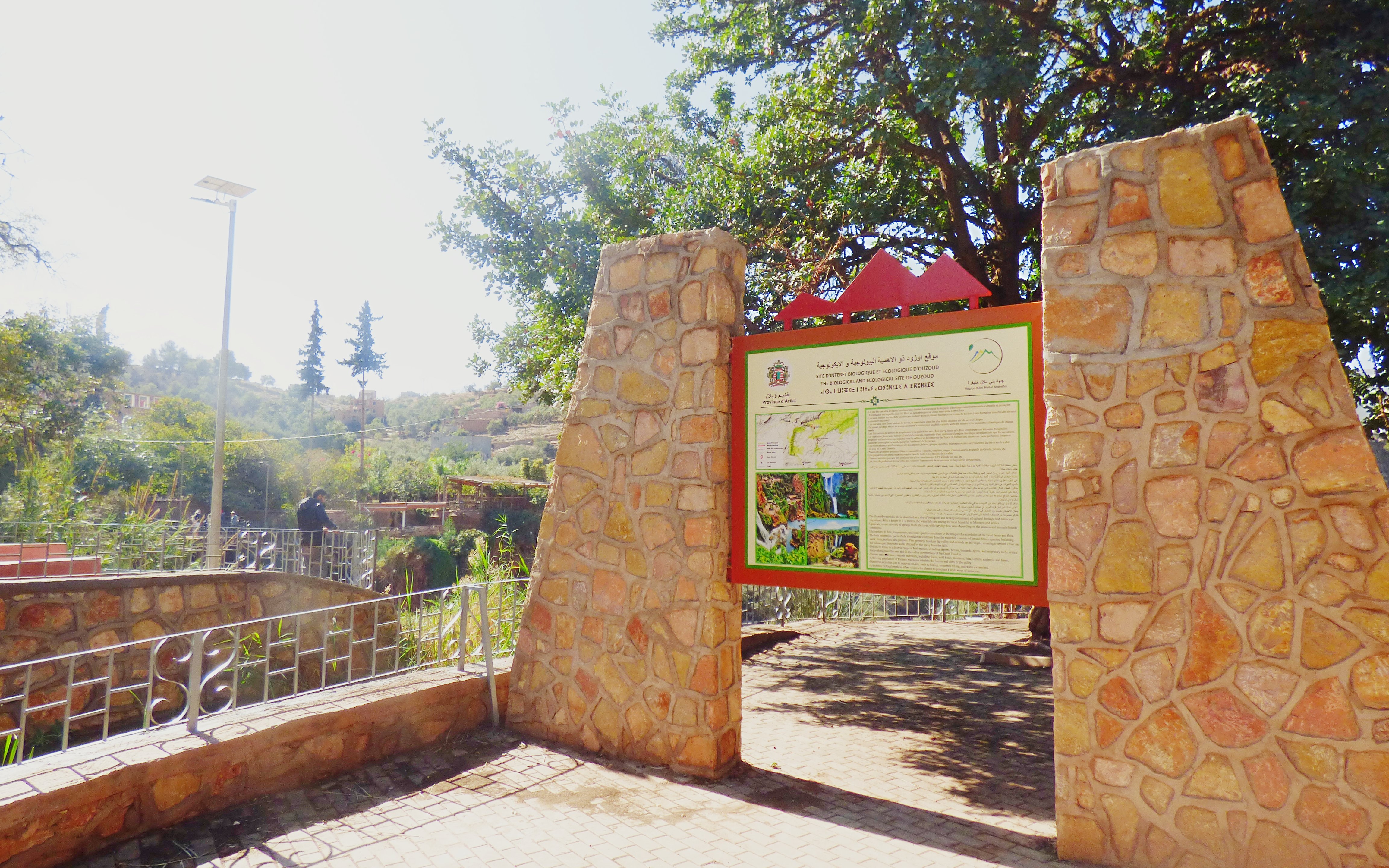 Entrance sign at Ouzoud Waterfalls trailhead, Morocco, with tourist in background.