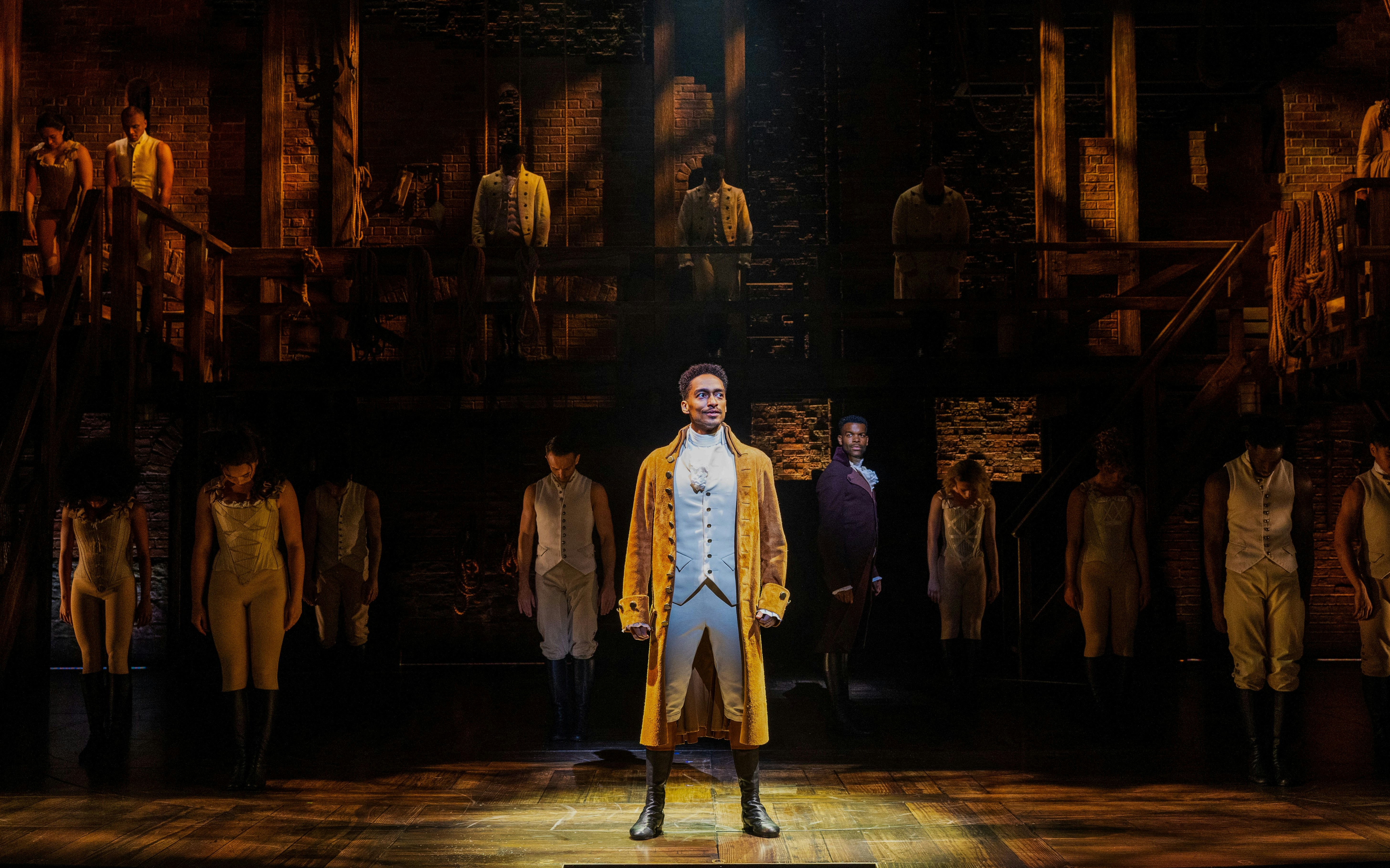 Cast performing on stage in Hamilton production, featuring lead actor in period costume.