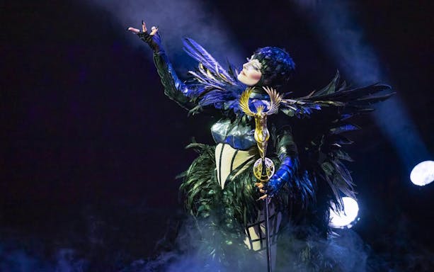 Performer in elaborate costume with feathers and staff during "Awakening" show.