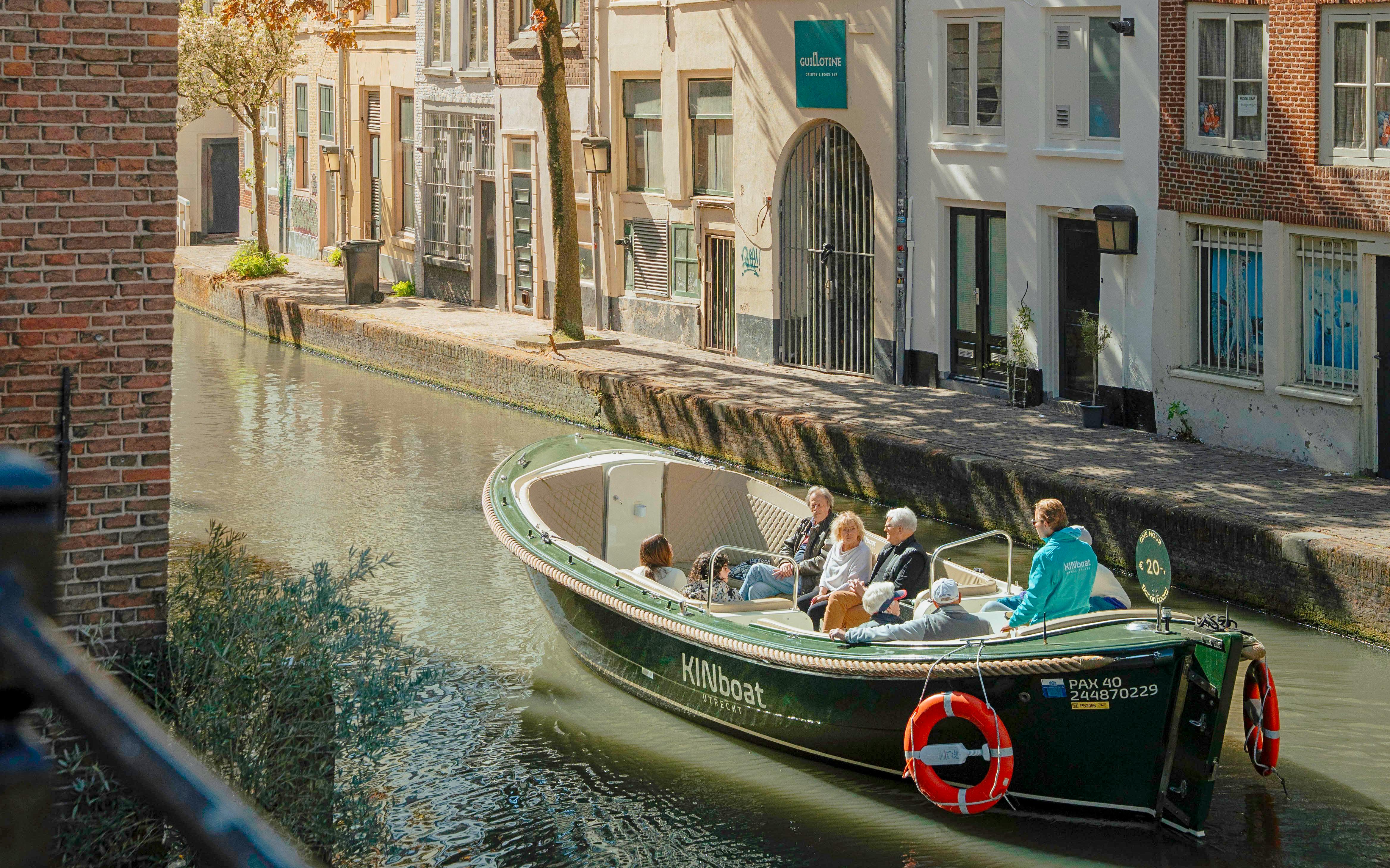 Open boat with tourists cruising a canal in Utrecht, passing historic buildings.