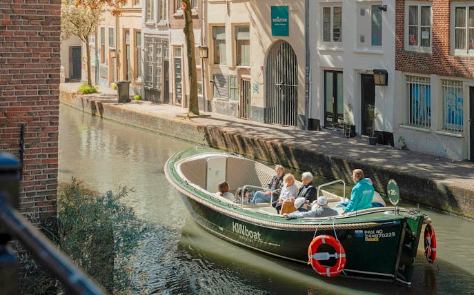 Open boat with tourists cruising a canal in Utrecht, passing historic buildings.