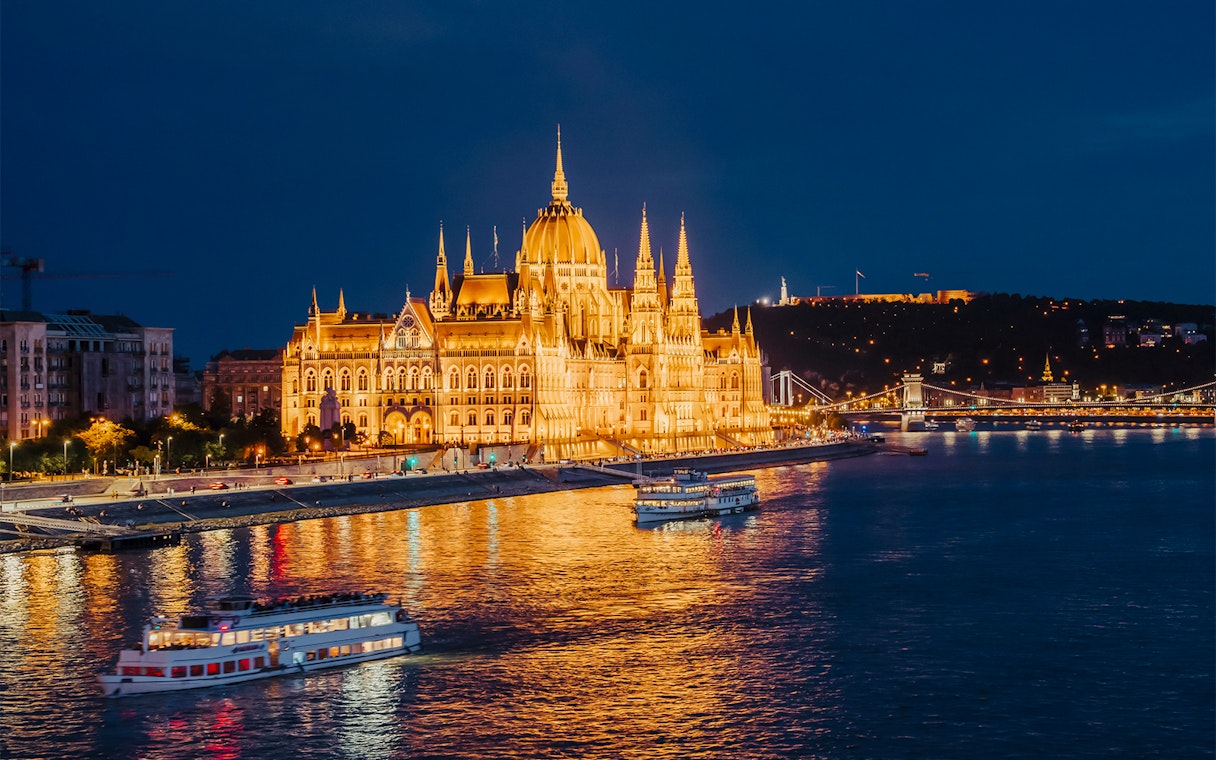 Budapest Parliament illuminated at night with cruise boats on the Danube River.