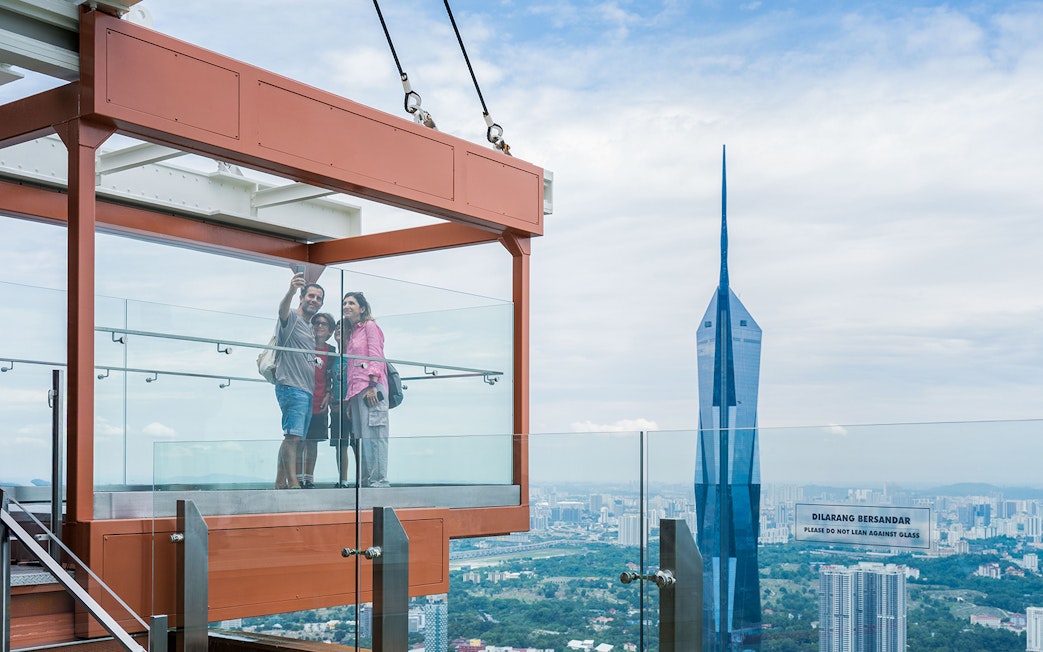 Family taking a selfie in the glass box at Kuala Lumpur Tower with city skyline view.