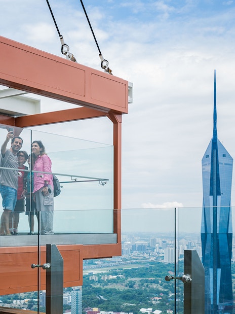 Family taking a selfie in the glass box at Kuala Lumpur Tower with city skyline view.