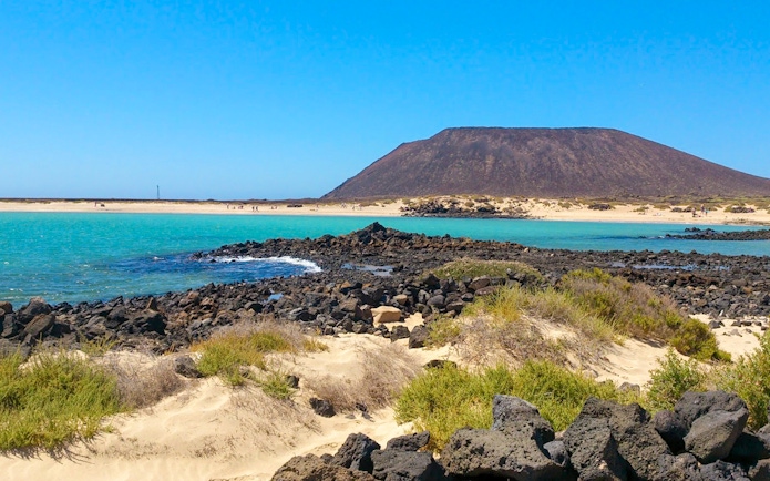 Rocky shoreline and turquoise waters of Lobos Island with volcanic landscape in the background.