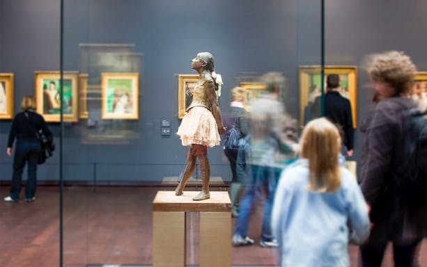 Sculpture of a ballerina in Orsay Museum, Paris, with visitors viewing Impressionist paintings.