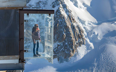 Person standing on a glass platform overlooking snowy peaks in Chamonix Mont Blanc.