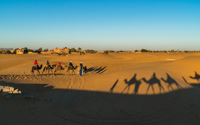 Camel caravan crossing Sahara desert near village.