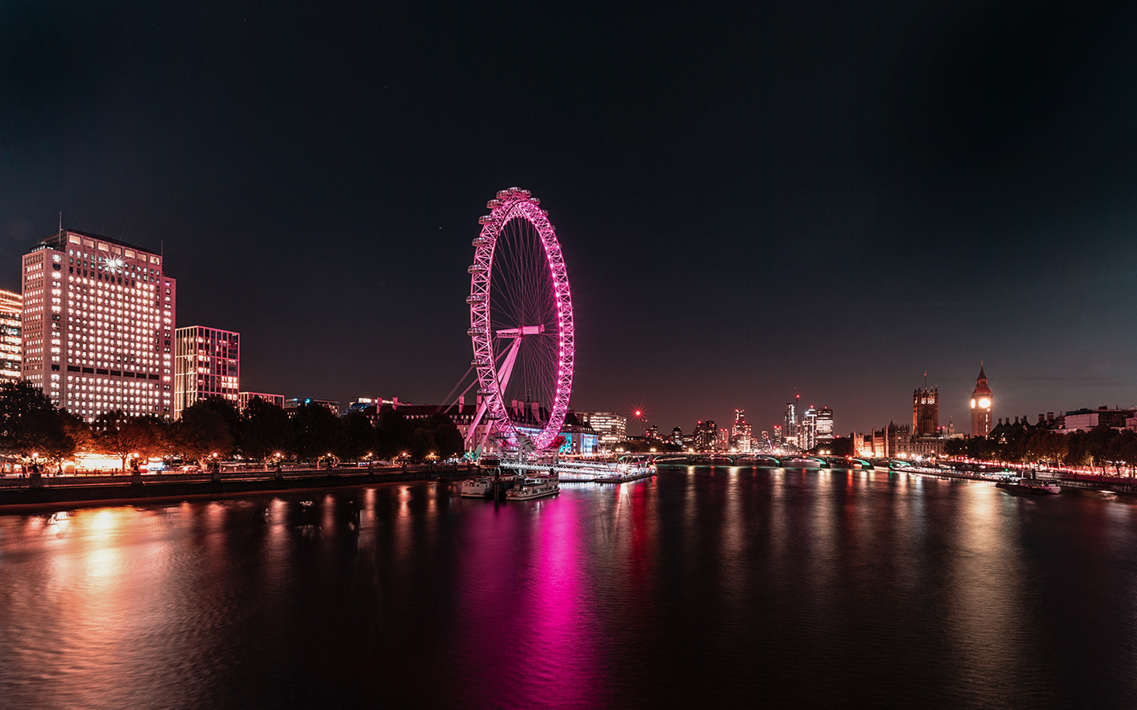 London Eye illuminated at night along the Thames River, with city skyline in the background.