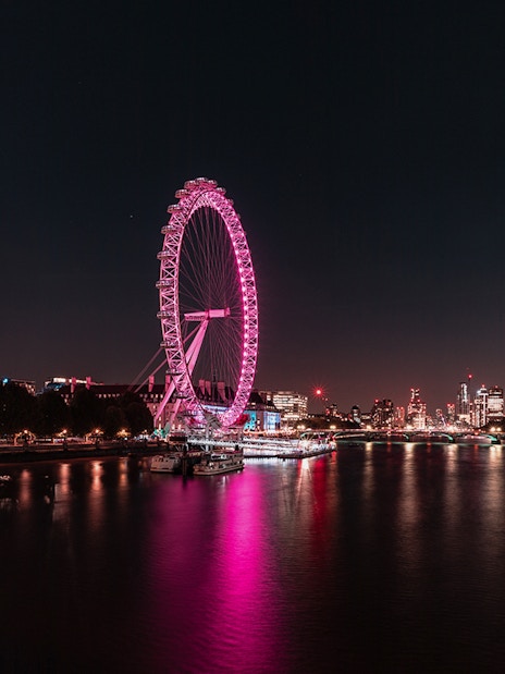 London Eye illuminated at night along the Thames River, with city skyline in the background.