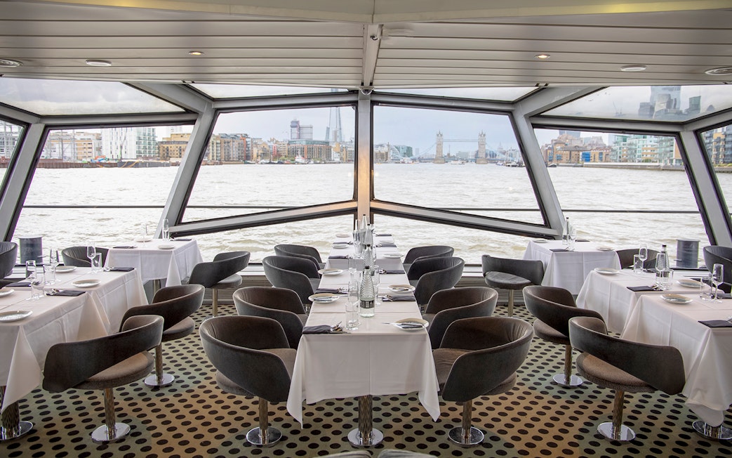 Dining area with tables set for a meal on a cruise ship, view of Tower Bridge and London skyline.