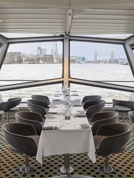 Dining area with tables set for a meal on a cruise ship, view of Tower Bridge and London skyline.