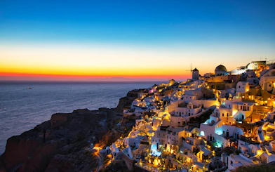 Oia village at sunset, Santorini, with illuminated white buildings and windmill overlooking the sea.