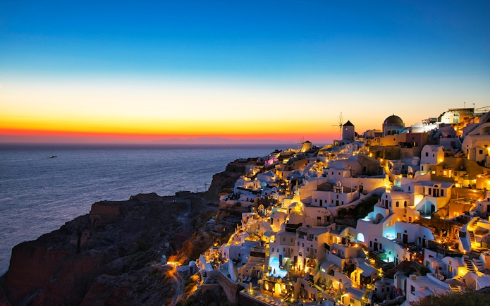 Oia village at sunset, Santorini, with illuminated white buildings and windmill overlooking the sea.