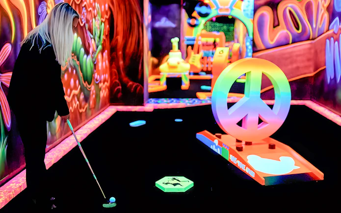 Person playing blacklight minigolf at Rock of Ages with neon peace sign obstacle.