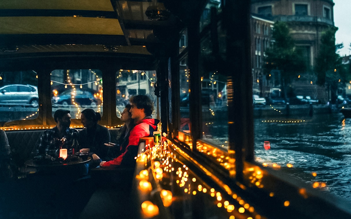 Cozy canal cruise in Amsterdam with people enjoying drinks and snacks, surrounded by festive lights.