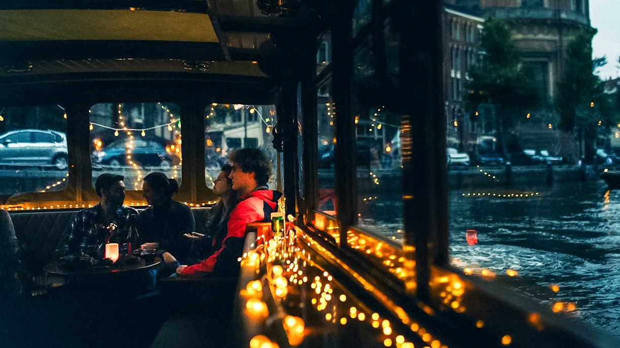 Cozy canal cruise in Amsterdam with people enjoying drinks and snacks, surrounded by festive lights.