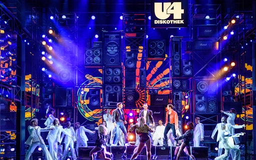 Performers on stage at Ronacher for "Rock Me Amadeus – The Falco Musical" with vibrant lights and speakers.