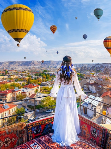 Hot air balloons over Goreme Valley with a person in traditional attire on a rooftop.