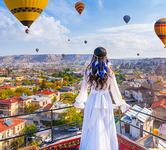 Hot air balloons over Goreme Valley with a person in traditional attire on a rooftop.