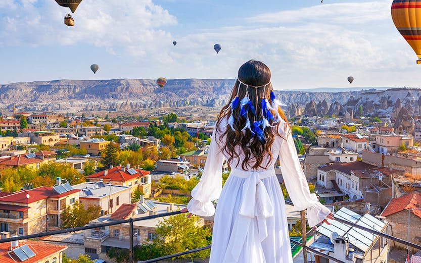 Hot air balloons over Goreme Valley with a person in traditional attire on a rooftop.