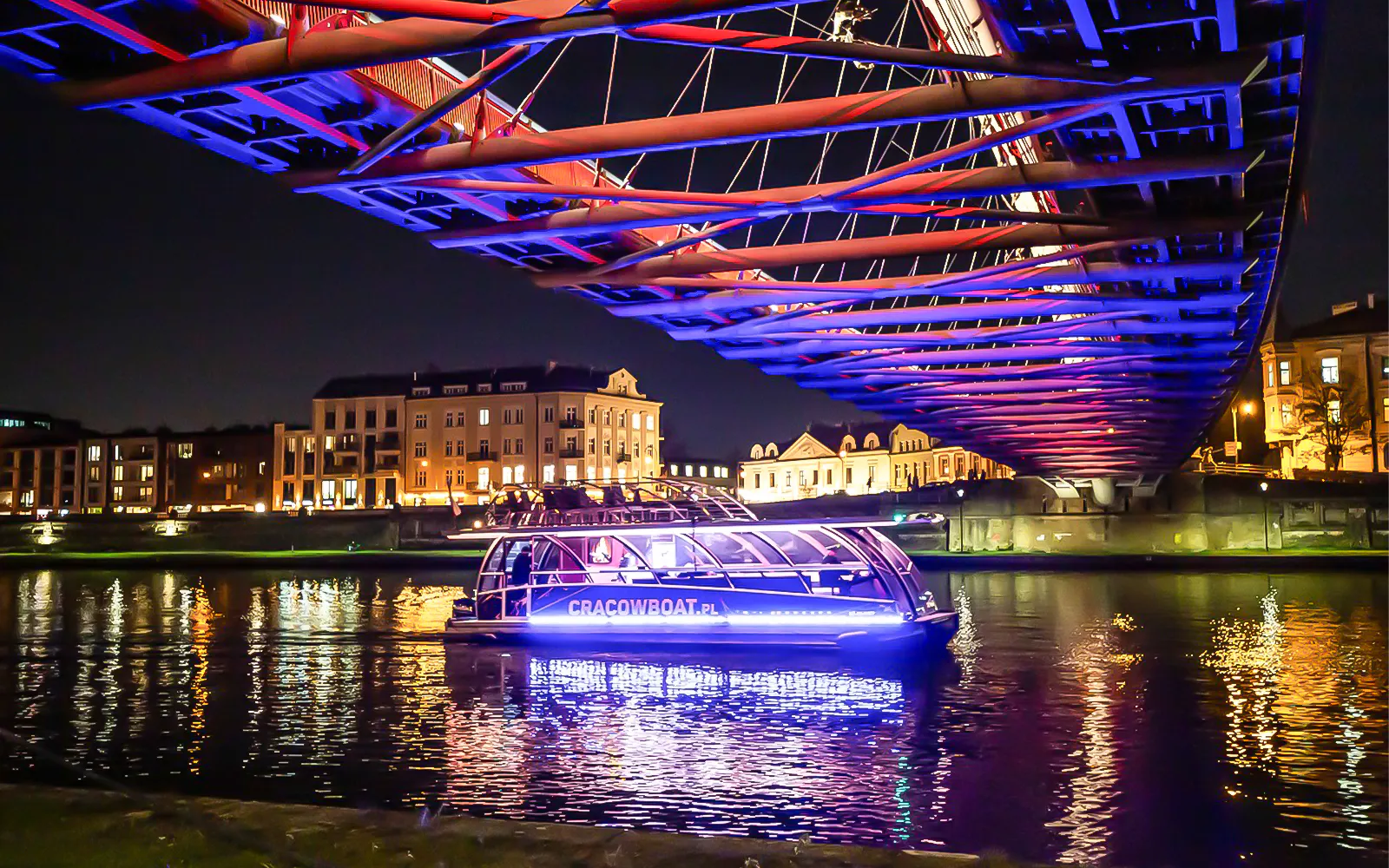 Catamaran on Vistula River at night under illuminated bridge in Krakow.
