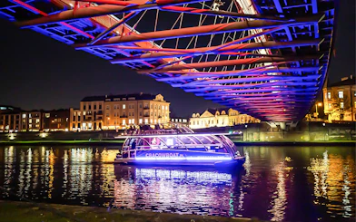 Catamaran on Vistula River at night under illuminated bridge in Krakow.