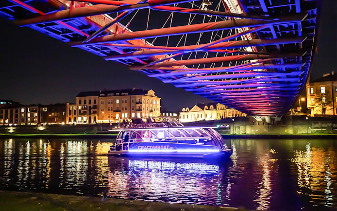 Catamaran on Vistula River at night under illuminated bridge in Krakow.